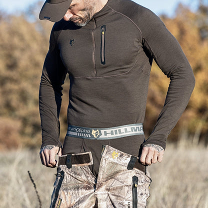 A person in a dark long-sleeve shirt adjusts their Hillman 2BL Merino Wool Hunting Long Johns outdoors, with a blurred backdrop of trees and grass.