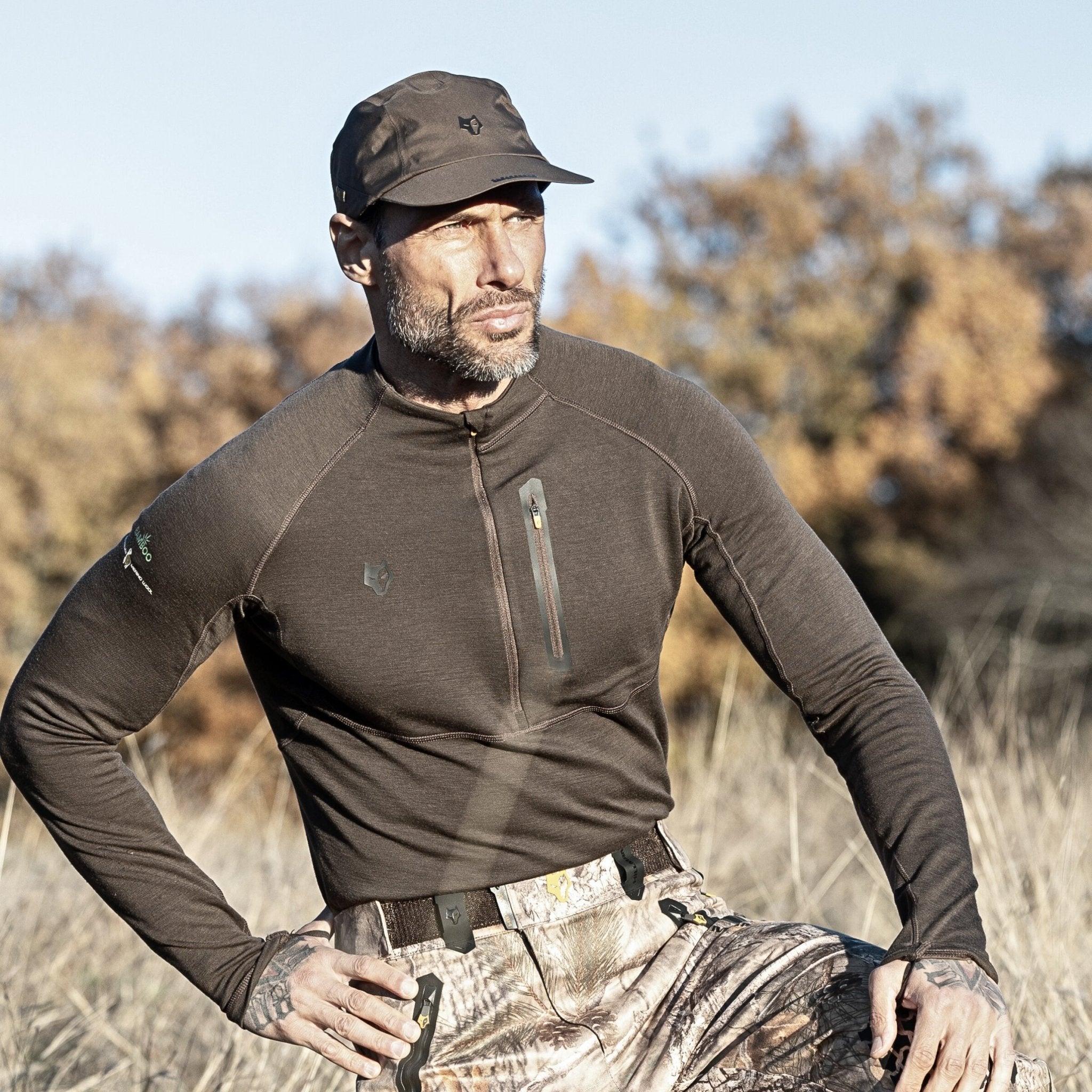 A man in a grassy field with dry vegetation and trees, wearing Hillmans 2BL Merino Wool Base Layer-Hunting Long Sleeve Thermal T-shirt and camouflage pants, gazes into the distance.