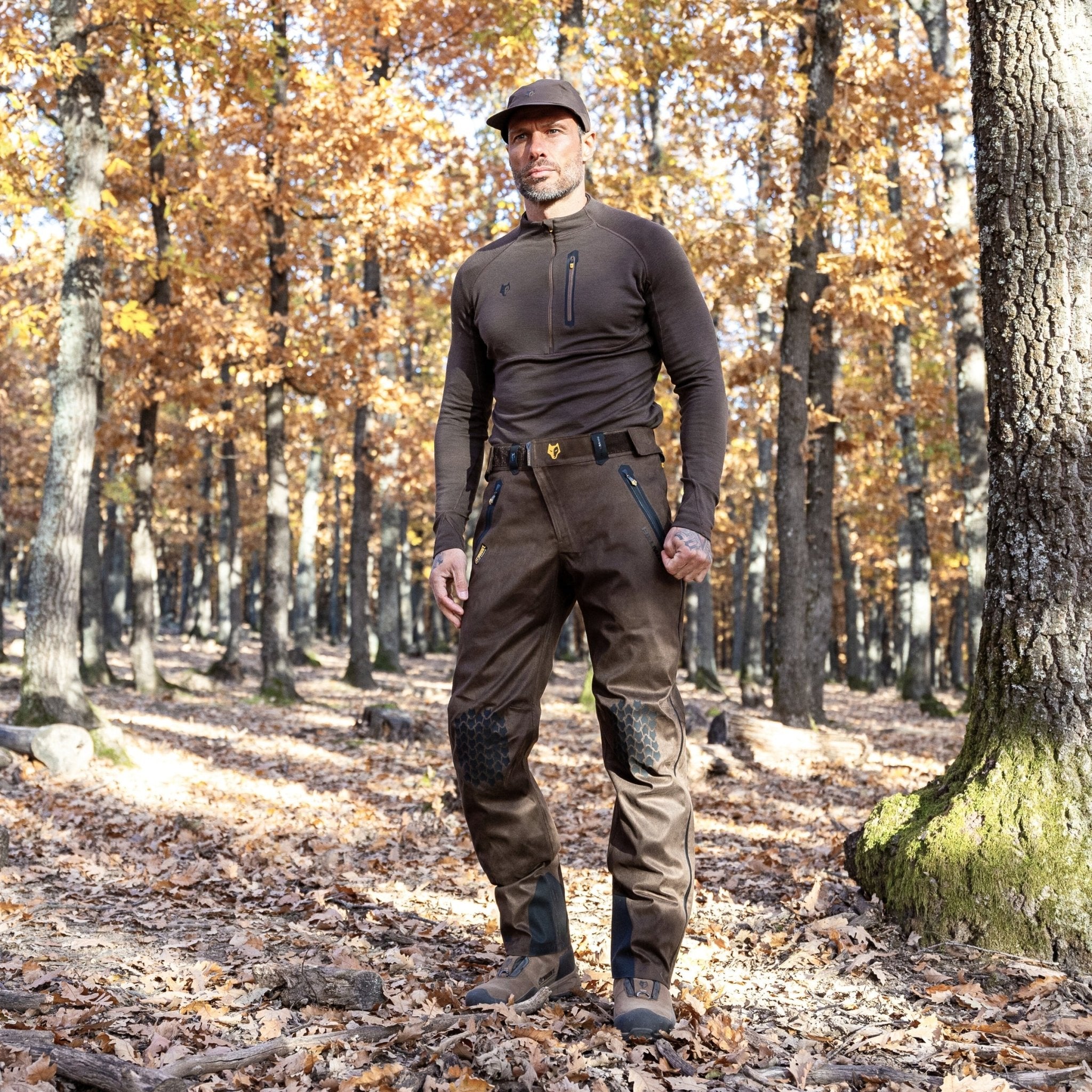 In an autumn forest with tall trees and orange leaves, a man in dark clothing confidently stands. Hes wearing a cap, Hillman 2BL Merino Wool Base Layer-Hunting Long Sleeve Thermal T-shirt, and utility pants with knee patches. The ground is blanketed in fallen leaves as he looks ahead.